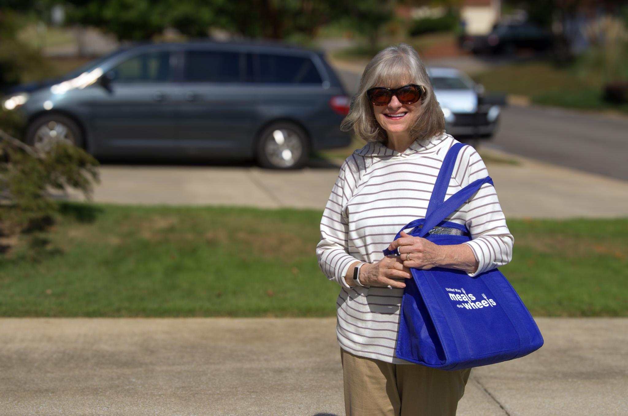 Meals on Wheels volunteer smiles while delivering a meal, holding a branded blue bag in front of a parked minivan.